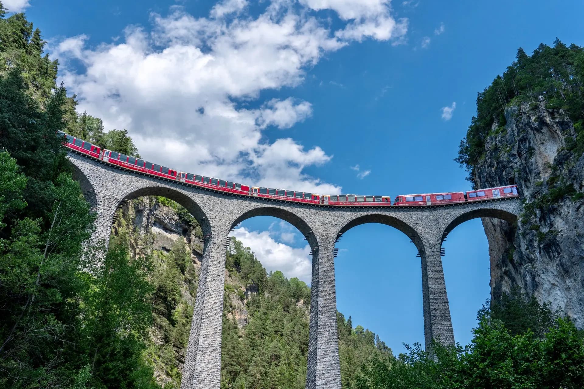 Tren rojo cruzando un gran viaducto de piedra bajo un cielo azul