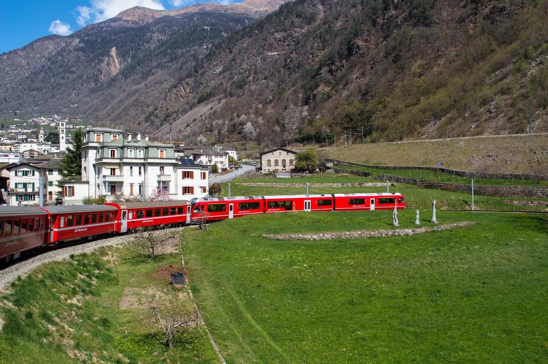 Tren rojo atravesando un valle alpino verde junto a un pequeño núcleo urbano
