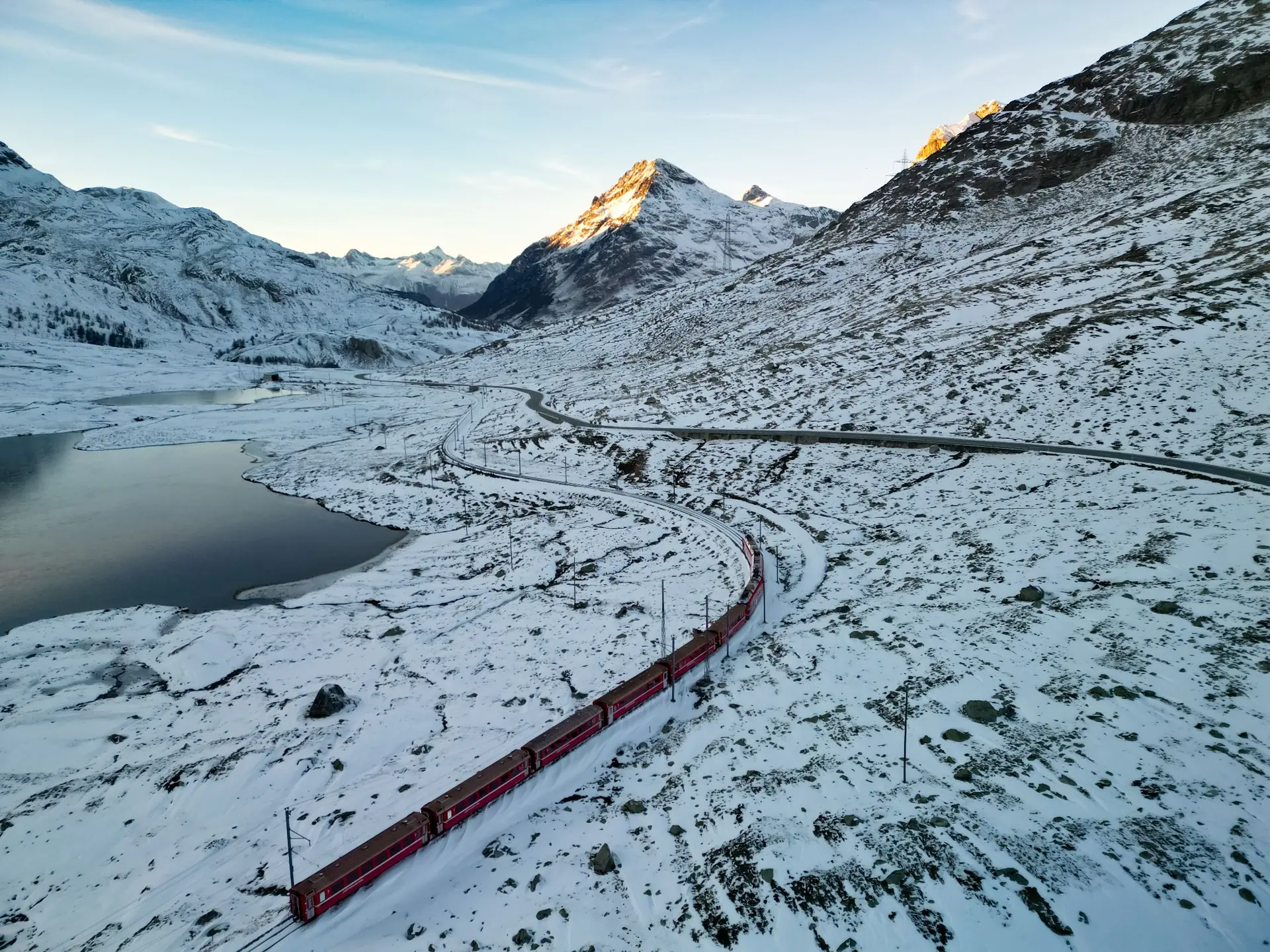 Tren rojo serpenteando entre lagos y montañas nevadas al amanecer